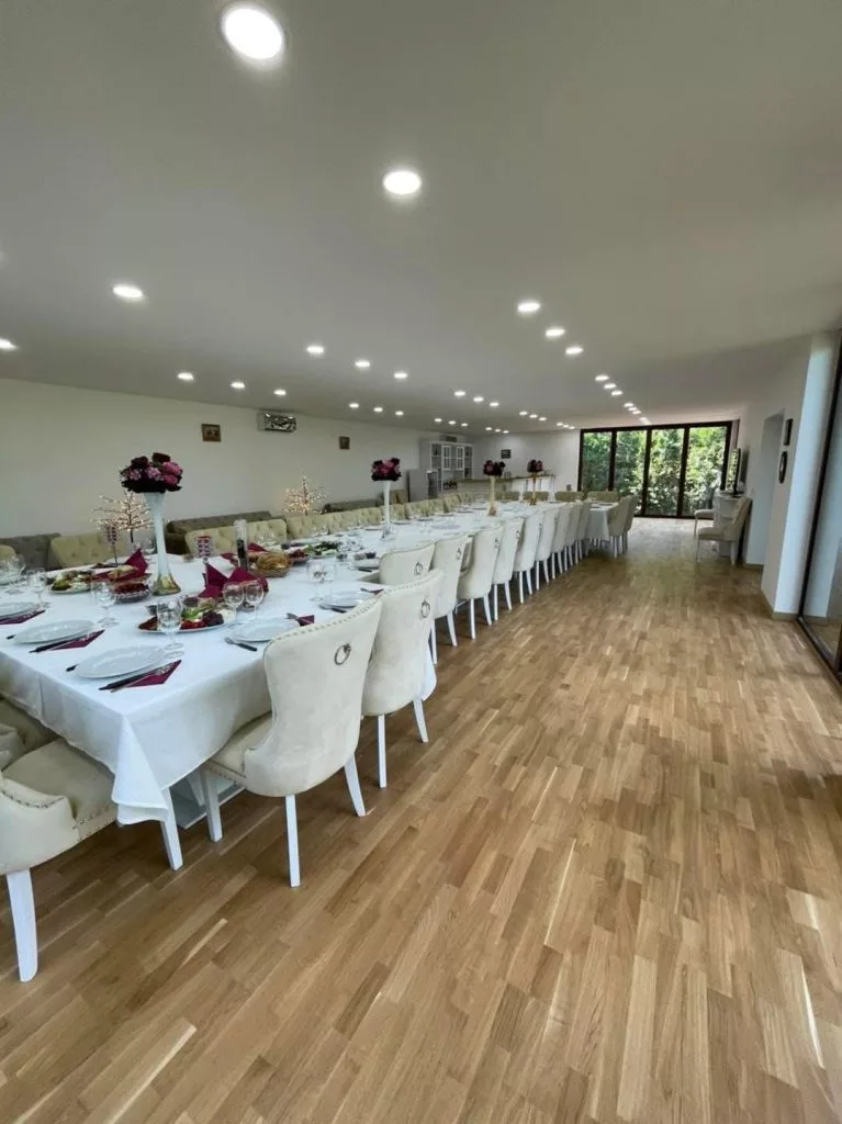Masă lungă aranjată festiv cu preparate, fețe de masă albe și scaune crem, într-o sală luminoasă