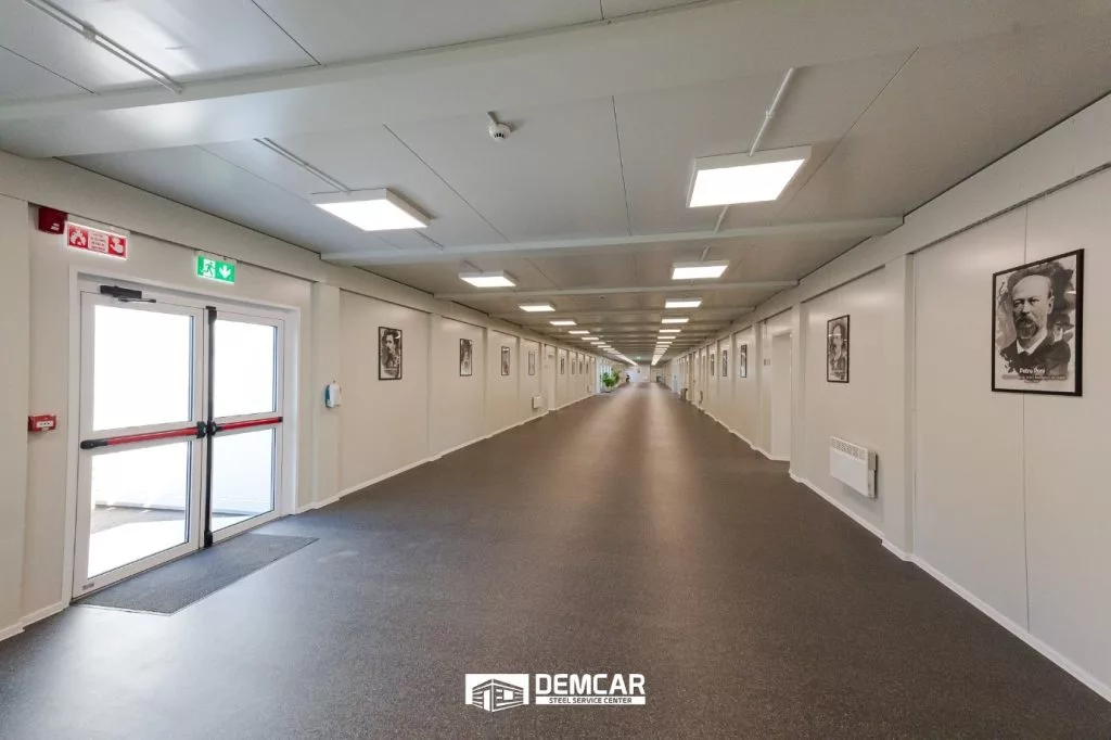 Coridor lung cu pardoseală gri, tablouri pe pereți și ușă de evacuare într-o clădire modulară